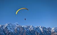The entire bottom third of the picture is covered by blue-tinged, snow capped mountains. The mountains are a dark brown, with a blue tinge. The upper part of the picture is of a clear blue sky. In the left center of the picture is a person parachuting ove