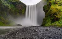 The top two thirds of the photo are of a moss covered, rocky cliff. It is cut in half, in the middle by a waterfall that runs down the left center of the picture and continues onto the bottom left of the picture. The bottom third of the picture is of a ro
